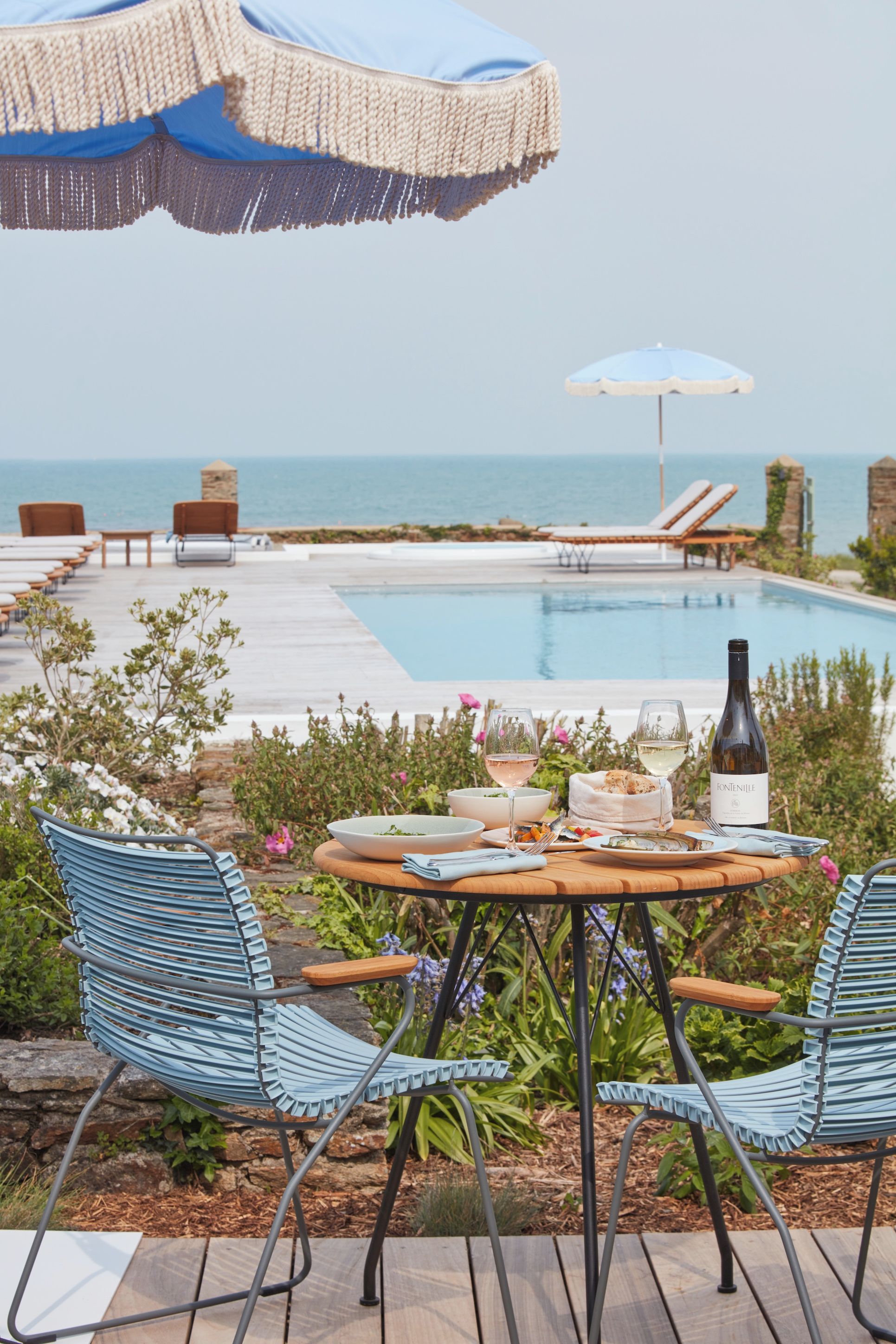 A poolside dining area with a table set for two, a bottle of wine, and a view of the sea.