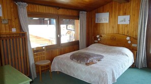 A room with a wooden headboard bed, a small wooden chair, a table, and a window with curtains.