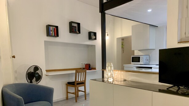 Ein modernes Wohnzimmer mit einem blauen Sessel, einem Holzhocker, einem weißen Schrank, einem Fernseher und einem kleinen Tisch mit Büchern und einer Lampe.