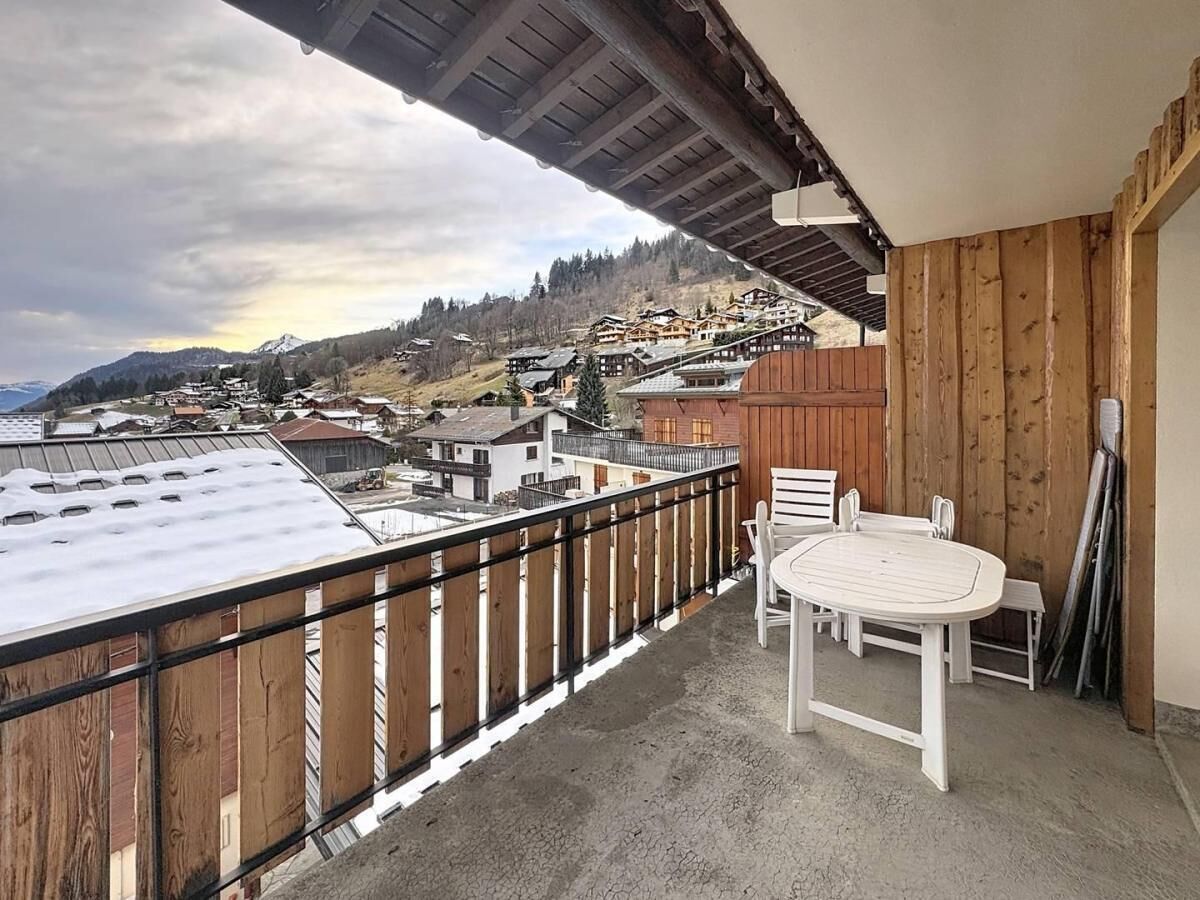 A balcony with a view of a mountainous village, wooden railings, and a table with chairs.