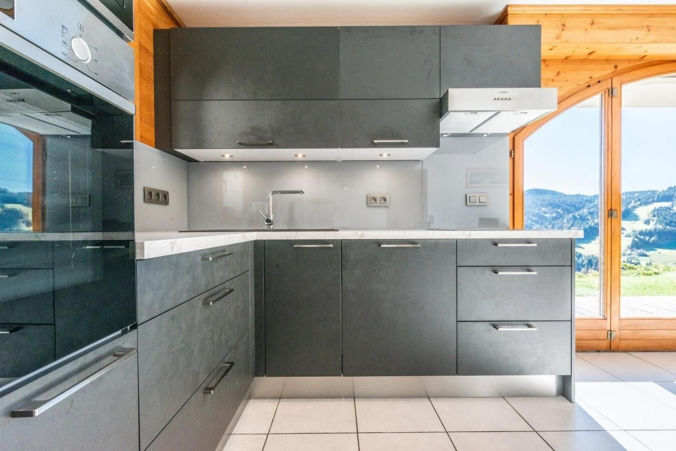 A modern kitchen with dark cabinetry, a stainless steel range hood, and a large window offering a view of the outdoors.
