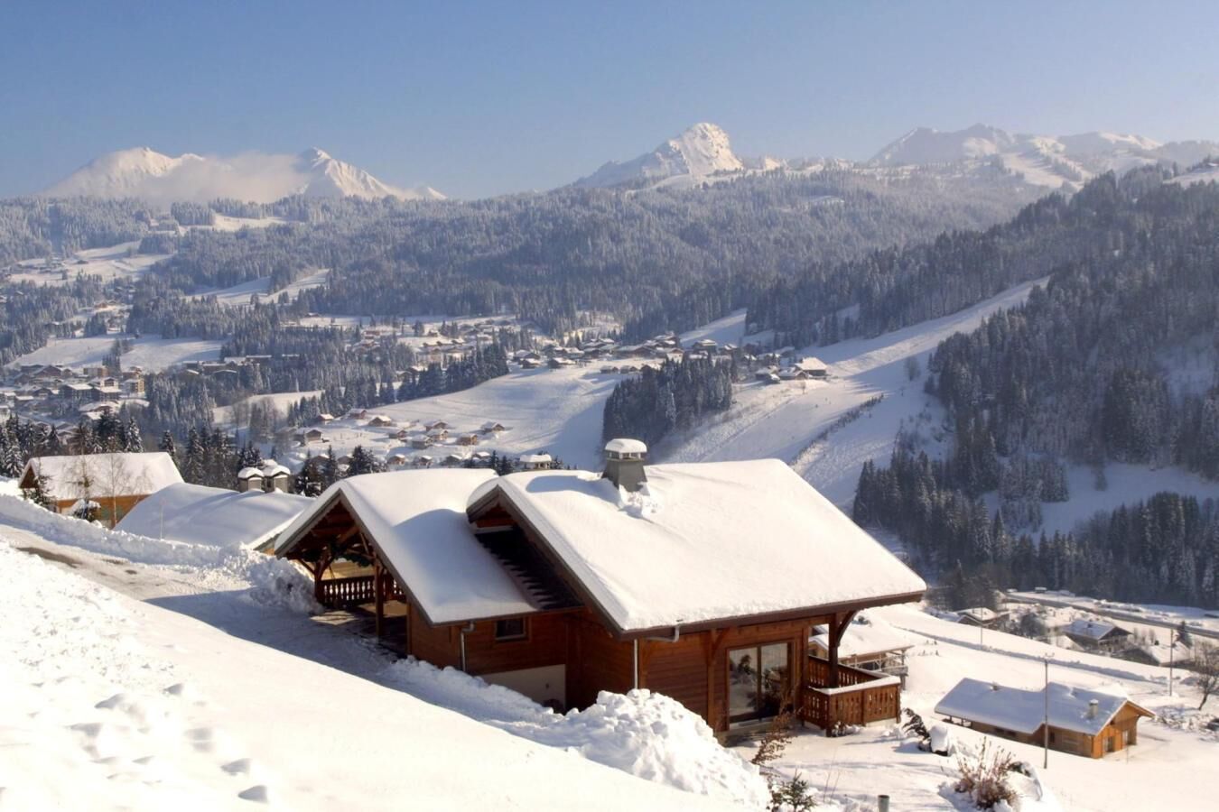 Una cabaña de madera con techo nevado, terraza de madera y vista a un paisaje montañoso cubierto de nieve.