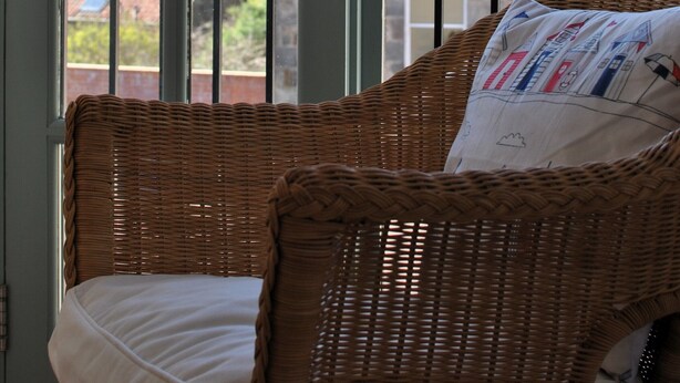 Una silla de mimbre con un cojín y una almohada con diseño estampado, ubicada frente a un ventanal con vista a un edificio de ladrillo.