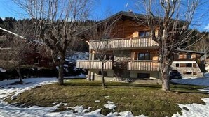 Un chalet en bois à deux étages, doté d’un balcon, entouré d’un paysage enneigé et d’arbres sans feuilles.