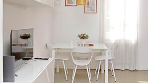 Un espace repas moderne avec une table blanche, quatre chaises blanches, une table avec un plateau en verre, un ordinateur portable, une télécommande et un vase rempli de fleurs.
