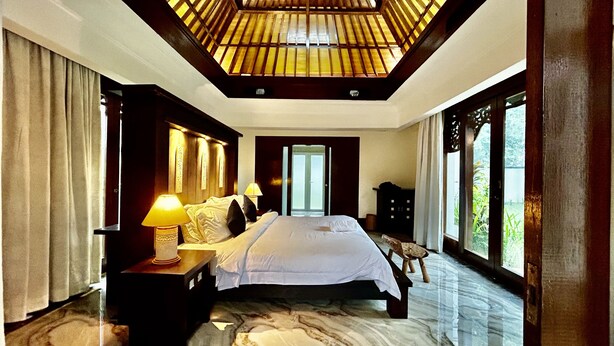 A spacious bedroom with a large bed, a nightstand with a lamp, and a view of greenery through the window. - Pat-Mase Villas Jimbaran (badung)