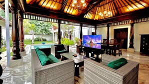 A covered outdoor seating area with wicker furniture, a television, and a pool in the background. - Pat-Mase Villas Jimbaran (badung)