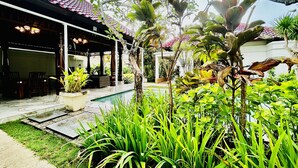 A covered outdoor area with a pool, surrounded by lush greenery and a building with a tiled roof. - Pat-Mase Villas Jimbaran (badung)