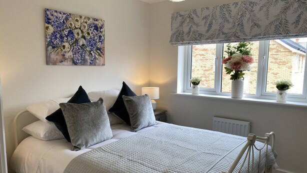 A bedroom with a bed, pillows, a bedside table, a lamp, a window with curtains, and a painting on the wall. - Comfortable Well Equipped Home In Isleham. Close to Newmarket, Ely & Cambridge (Isleham)