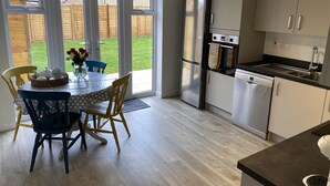 A modern kitchen with a refrigerator, oven, and dishwasher. There is a dining table with chairs and a view of a backyard through glass doors. - Comfortable Well Equipped Home In Isleham. Close to Newmarket, Ely & Cambridge (Isleham)