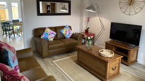 A living room with a brown leather sofa, a wooden coffee table, a TV on a wooden stand, a floor lamp, and a mirror on the wall. - Comfortable Well Equipped Home In Isleham. Close to Newmarket, Ely & Cambridge (Isleham)