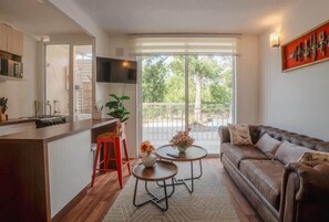 Una sala de estar moderna con un sofá de cuero marrón, una zona de cocina con un taburete rojo y un amplio ventanal con vistas a zonas verdes.
