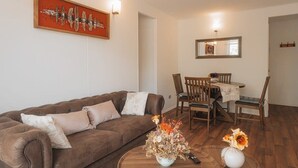 A living room with a brown sofa, a dining table with chairs, and a vase with flowers on a wooden coffee table. - Golden Pucon 3BR (Pucon)