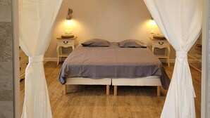 Un dormitorio con cama, cortinas blancas, dos mesitas de noche con lámparas y suelo de madera.