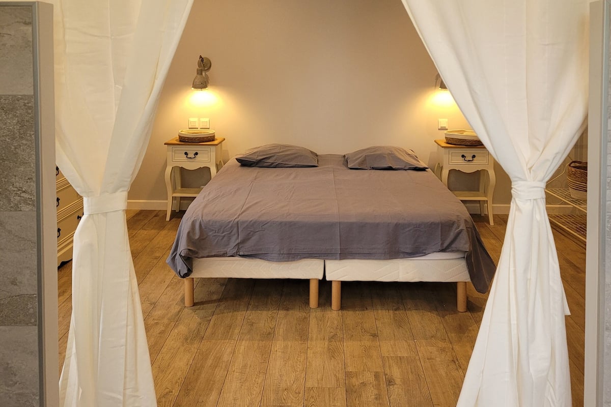 Ein Schlafzimmer mit einem Bett, weißen Vorhängen, zwei Nachttischen mit Lampen und Holzboden.