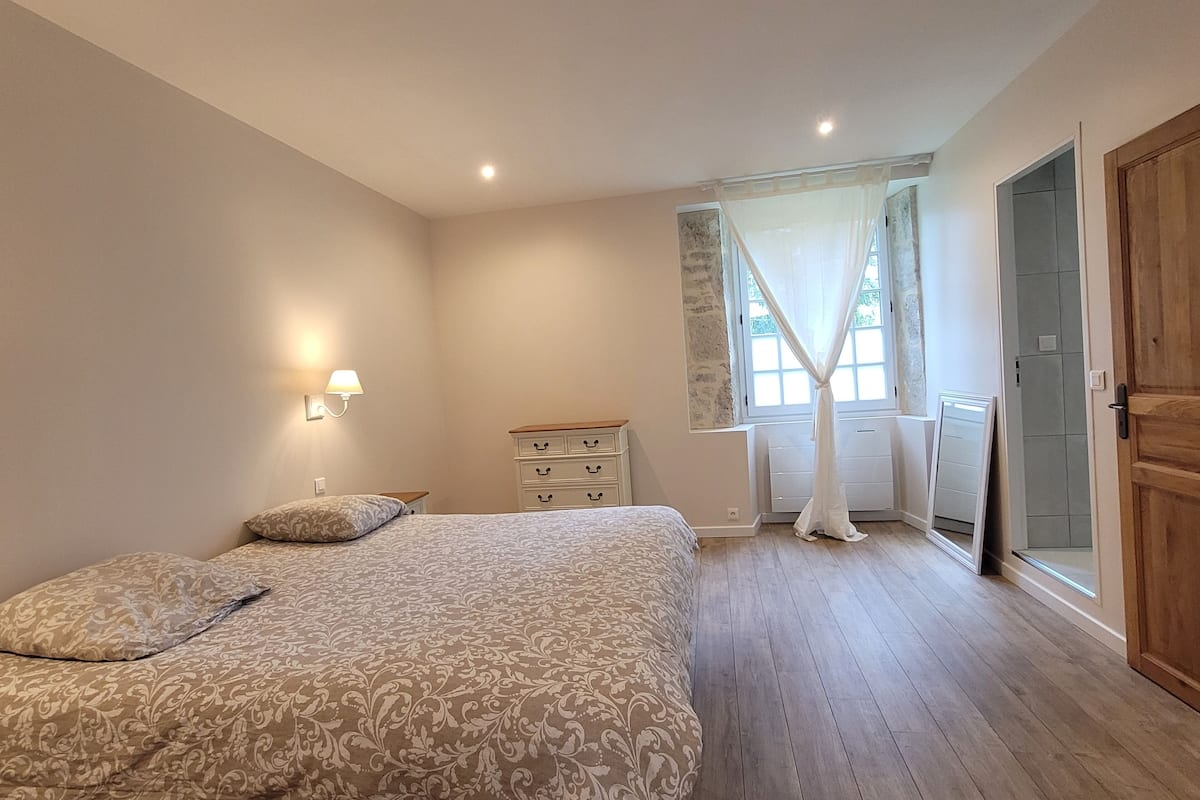 Ein Schlafzimmer mit Bett, Nachttisch, Lampe und einem Fenster mit Vorhängen.