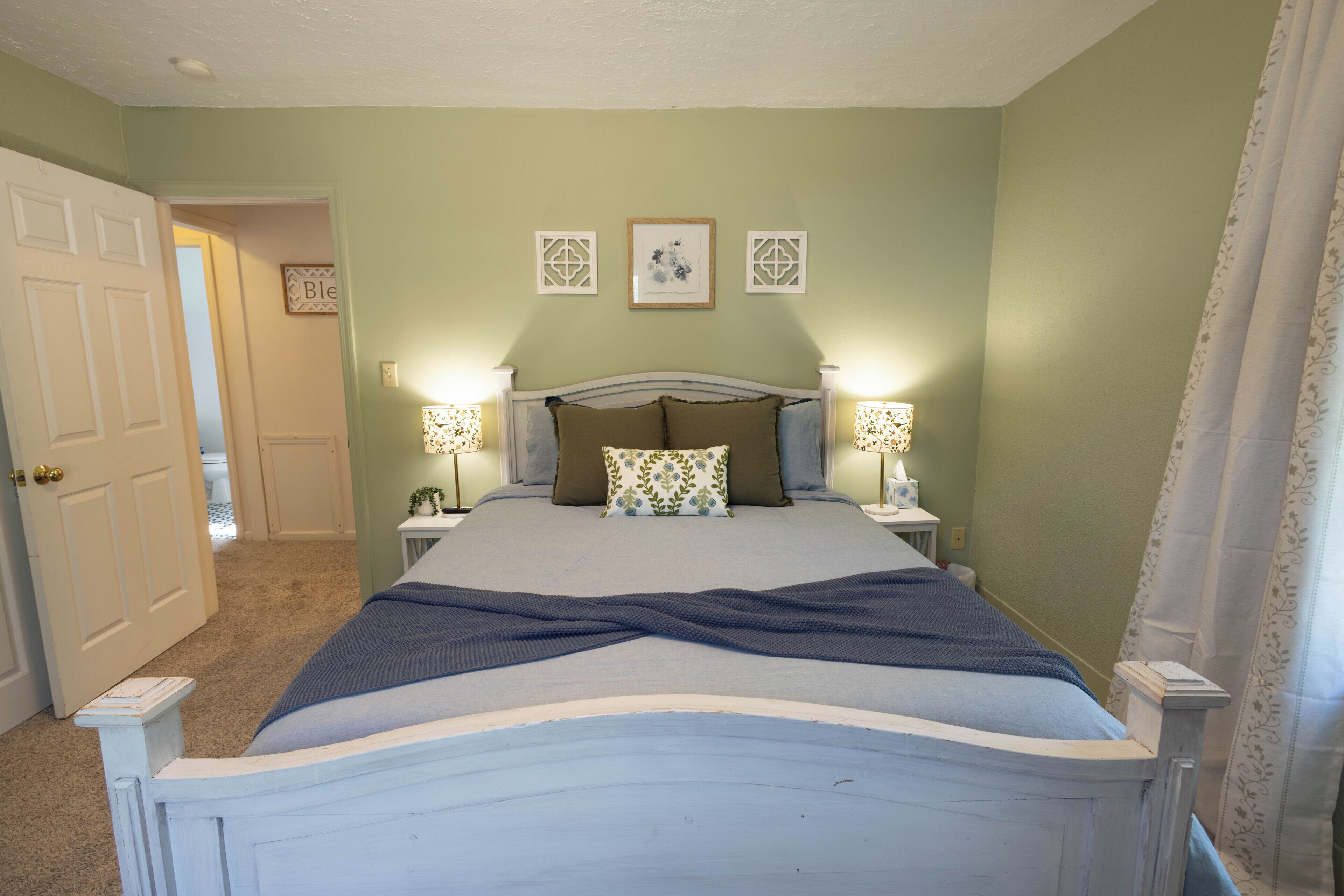 A bedroom with a bed, two bedside lamps, a door, and a wall with three framed pictures.