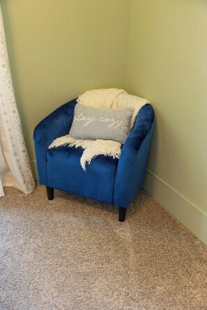 Un sillón de esquina con tela azul y un cojín gris, ubicado frente a una pared verde con una cortina blanca visible.