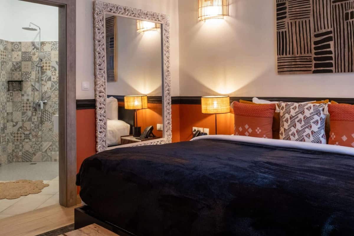 A modern hotel room with a large bed, a wooden bench, a shower area, and a large mirror.