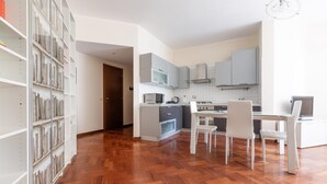 A modern kitchen with white cabinets, a microwave, and a stove. There are two white chairs and a dining table. The floor is wooden, and there is a bookshelf with books on the left. - Italianway Easy - Sismondi 74 (Milan)