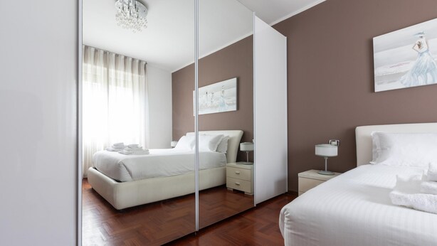 A modern hotel room with a large mirror, a bed, a bedside table, a lamp, and a painting on the wall. - Italianway Easy - Sismondi 74 (Milan)