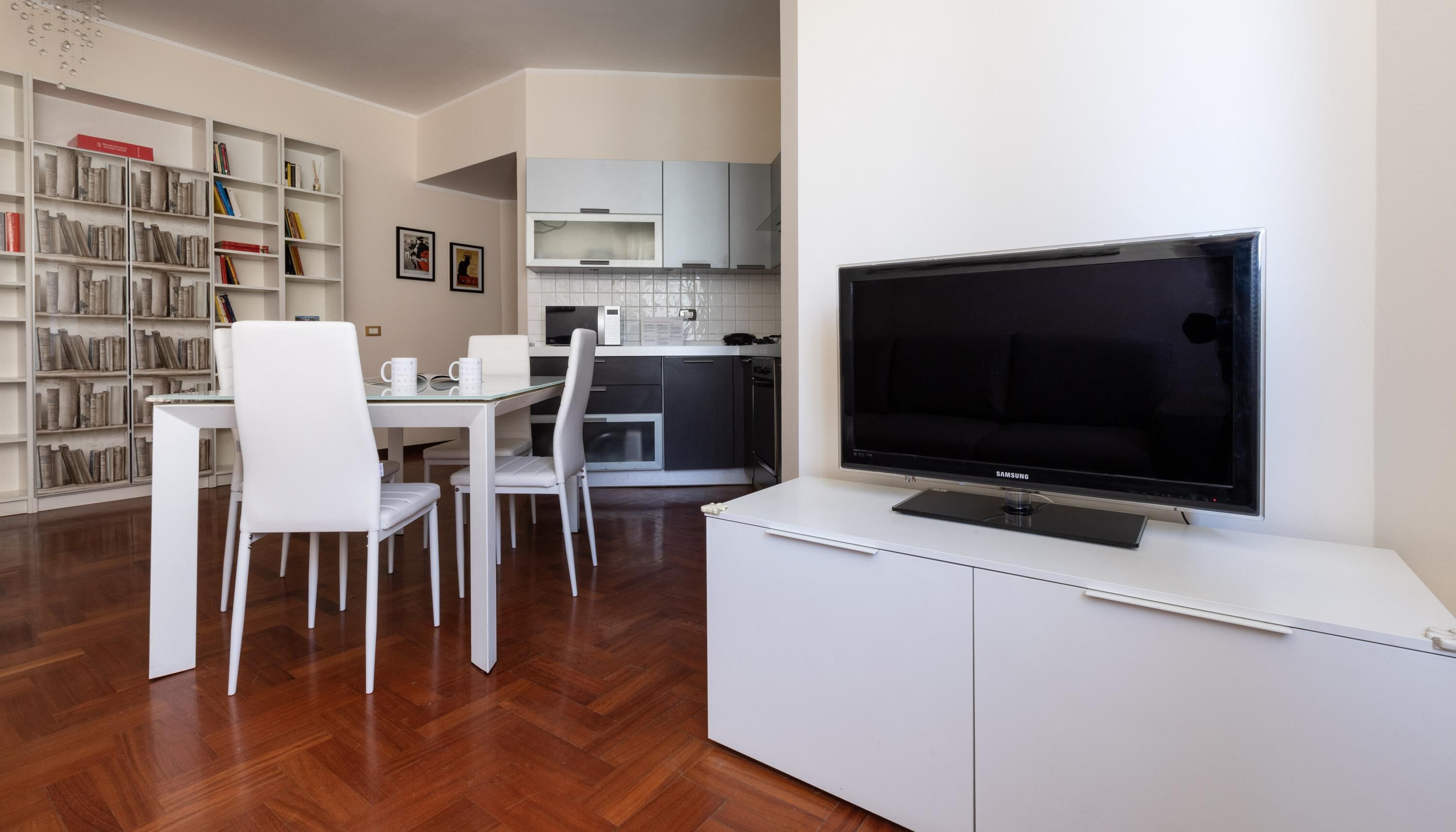 Una sala de estar moderna con televisor de pantalla plana, mobiliario blanco, una estantería y una zona de cocina.