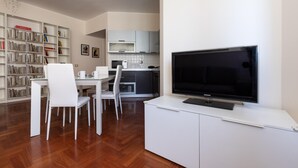 A modern living room with a flat-screen TV, white furniture, a bookshelf, and a kitchen area. - Italianway Easy - Sismondi 74 (Milan)