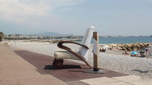 Een strandstoel met metalen frame en een witte kussens op een verhard pad, met een rotsachtige kustlijn en uitzicht op zee en verre gebouwen.