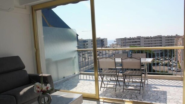 Een balkon met een bank, een kleine tafel met een bloevas, en uitzicht op gebouwen en een parkeerplaats.