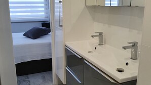 Een moderne badkamer met een witte wastafel, twee kranen en een spiegel erboven. In de aangrenzende kamer staat een bed met een zwart kussen.