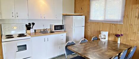 A compact kitchen with white appliances, a dining table with blue chairs, and a window with blinds.