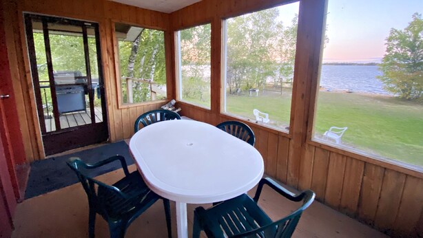 A covered outdoor dining area with a table and chairs, large windows offering views of a grassy area and a body of water. - "Shore Thing" Sunset Views (Seven Sisters Falls)