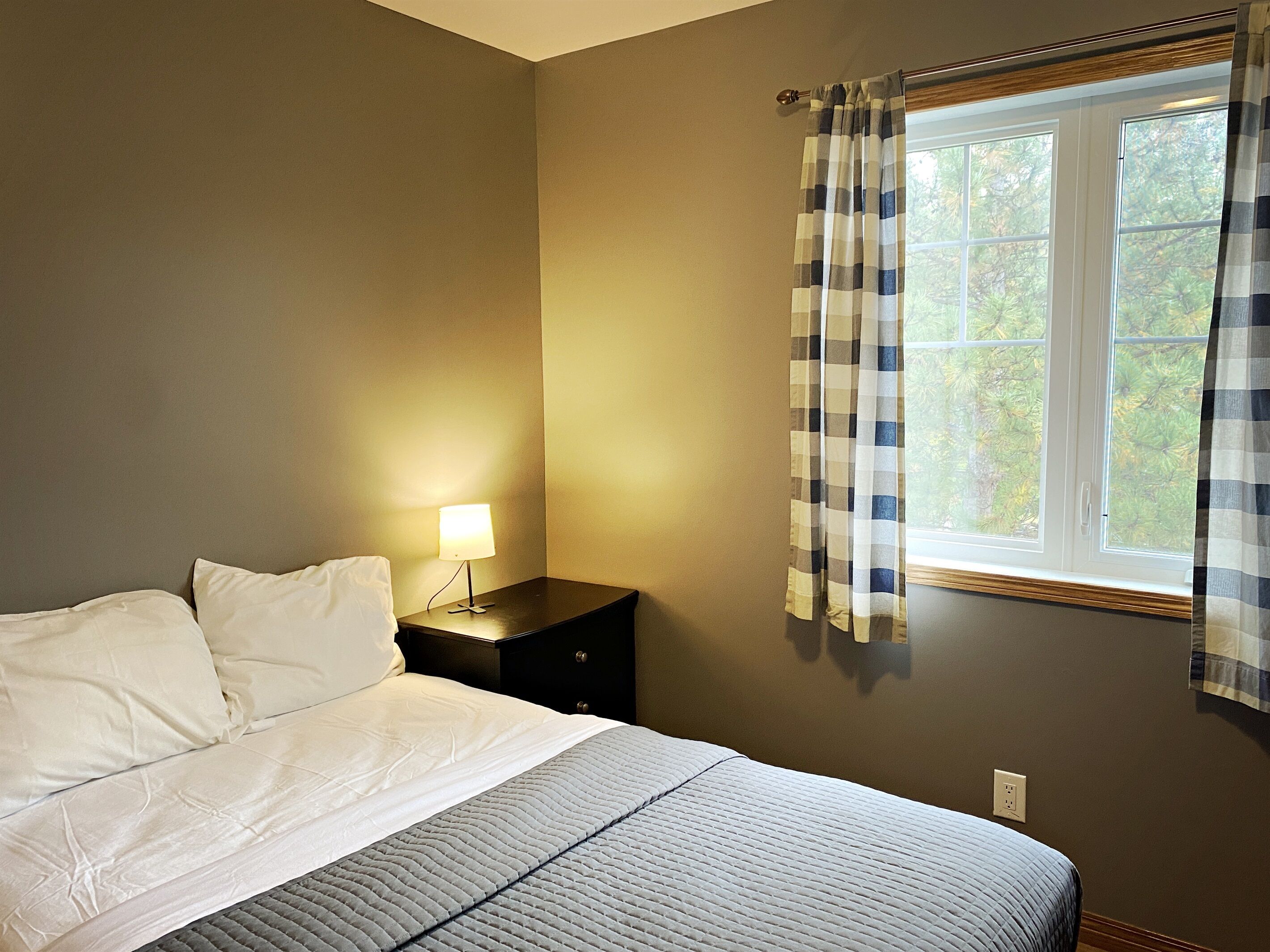 A neatly made bed with white linens and a gray quilt, a bedside table with a lamp, and a window with plaid curtains.