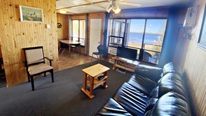 A cozy living room with a wooden wall, a black leather sofa, a wooden coffee table, a ceiling fan, and a view of the sea through large windows. - "Lakeside Landing" Escape to paradise (Seven Sister Falls)