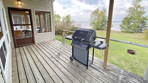 Een overdekt terras met een barbecue, een glazen deur naar een patio met buitenmeubilair en uitzicht op een meer.