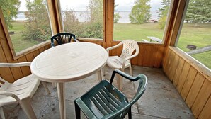Een overdekt buitengebied met een ronde tafel en stoelen, grote ramen met uitzicht op een gazon en bomen.