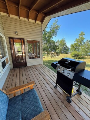 A covered outdoor deck with a grill, a door leading to a patio, and a view of the surrounding landscape.