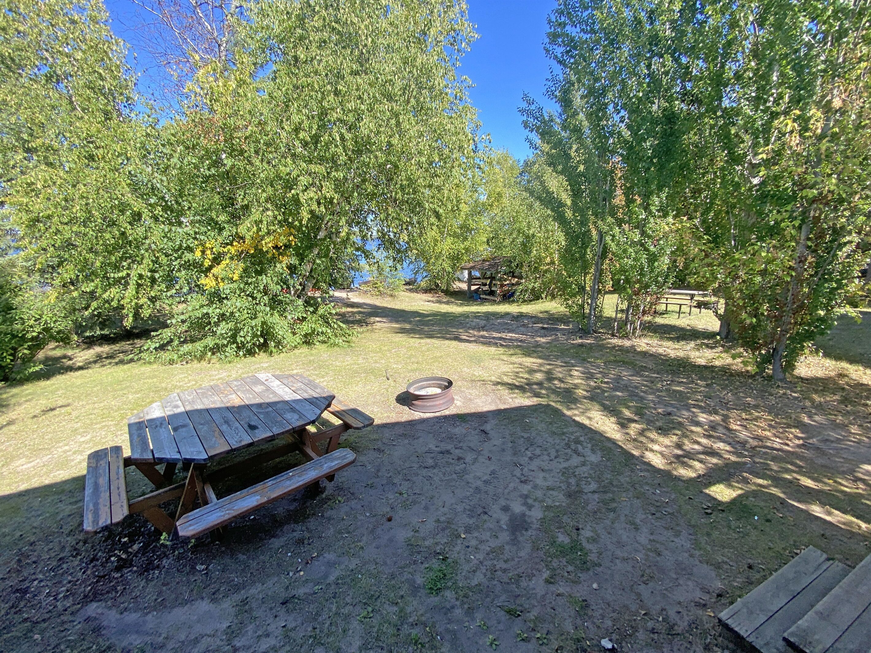 A picnic table with benches, a fire pit, and a wooden structure in a wooded area.