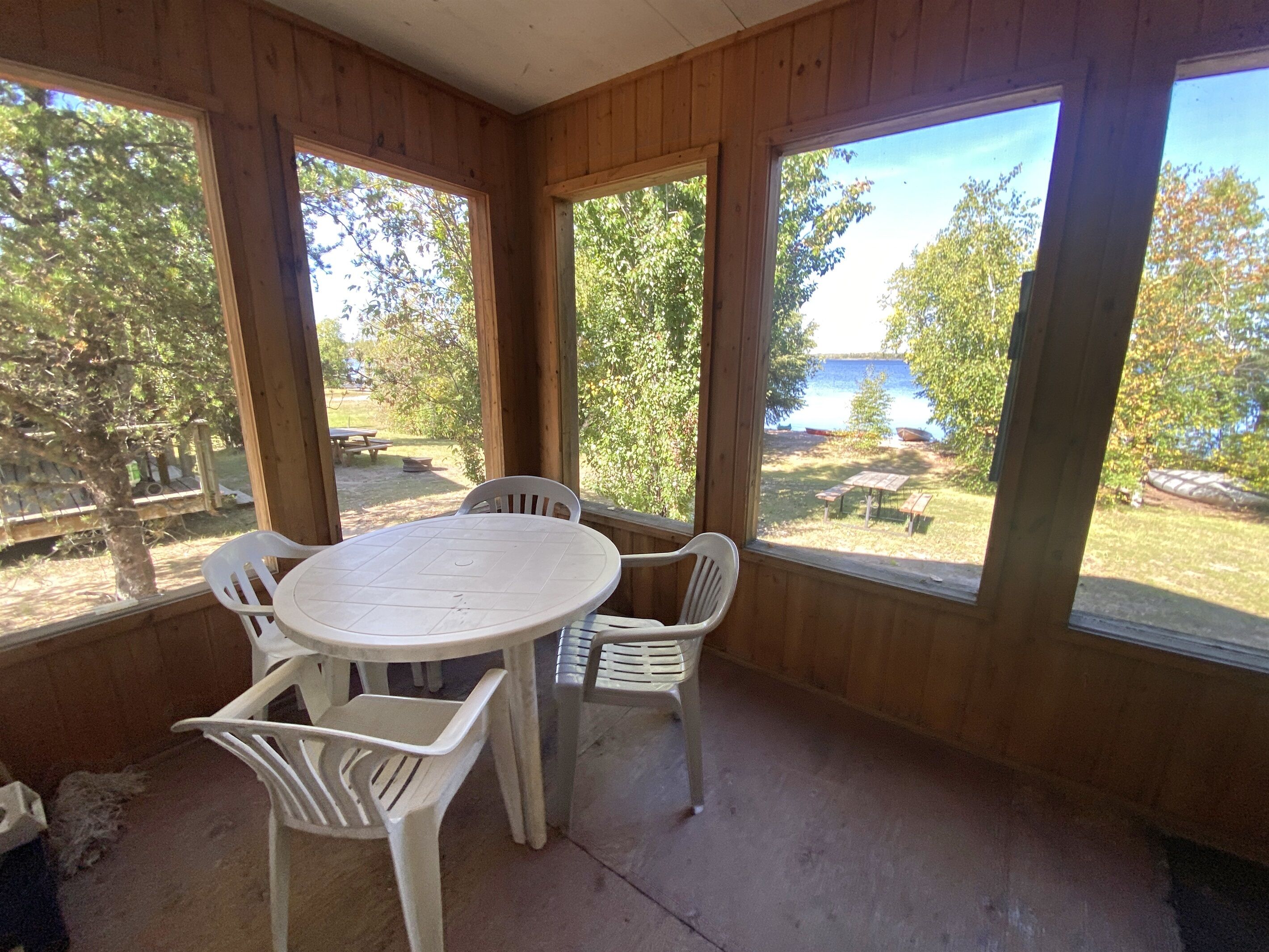 A wooden-paneled room with a round table and four white plastic chairs. Large windows offer views of a grassy area with picnic tables and trees.