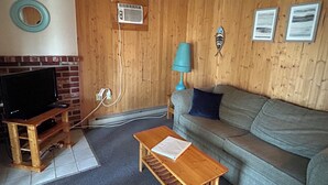 Uma sala de estar com televisão sobre um suporte de madeira, um sofá, uma pequena mesa de madeira e um ar condicionado fixado na parede.