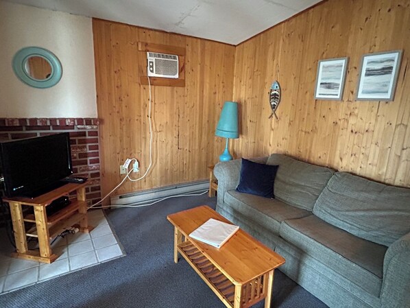 Ein Wohnzimmer mit Fernseher auf einem Holzständer, einer Couch, einem kleinen Holztisch und einer Wand-Klimaanlage.