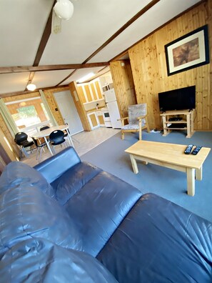 A room with a sofa, a wooden coffee table, a TV on a stand, and a kitchen area in the background. - "Family Tides" Enjoy our sandy beach! (Seven Sister Falls)