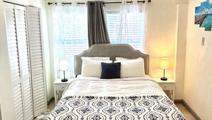 Un dormitorio con cama, ventilador de techo, mesita de noche con lámpara, ventana con cortinas y puerta con contraventanas.