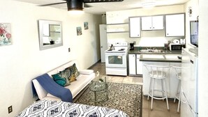 Un espacio habitable compacto con kitchenette, un sofá con cojines estampados, un ventilador de techo y una pequeña zona de comedor con taburetes de bar.