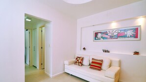 A white living room with a white sofa, a colorful abstract painting, and a small shelf with decorative items. - Quarto & Sala a 150m da Praia U052 (Rio de Janeiro)