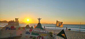 A beach scene with a sandcastle labeled "Feliz 2024 Rio de Janeiro 2024" and Brazilian flags. - Quarto & Sala a 150m da Praia U052 (Rio de Janeiro)