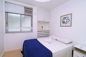 A bedroom with a bed, a window with blinds, a white wardrobe, a bedside table, and a framed picture on the wall. - Quarto & Sala a 150m da Praia U052 (Rio de Janeiro)