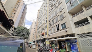 A multi-story building with balconies and air conditioning units, a street with parked motorcycles, and a visible storefront with a sign. - Quarto & Sala a 150m da Praia U052 (Rio de Janeiro)