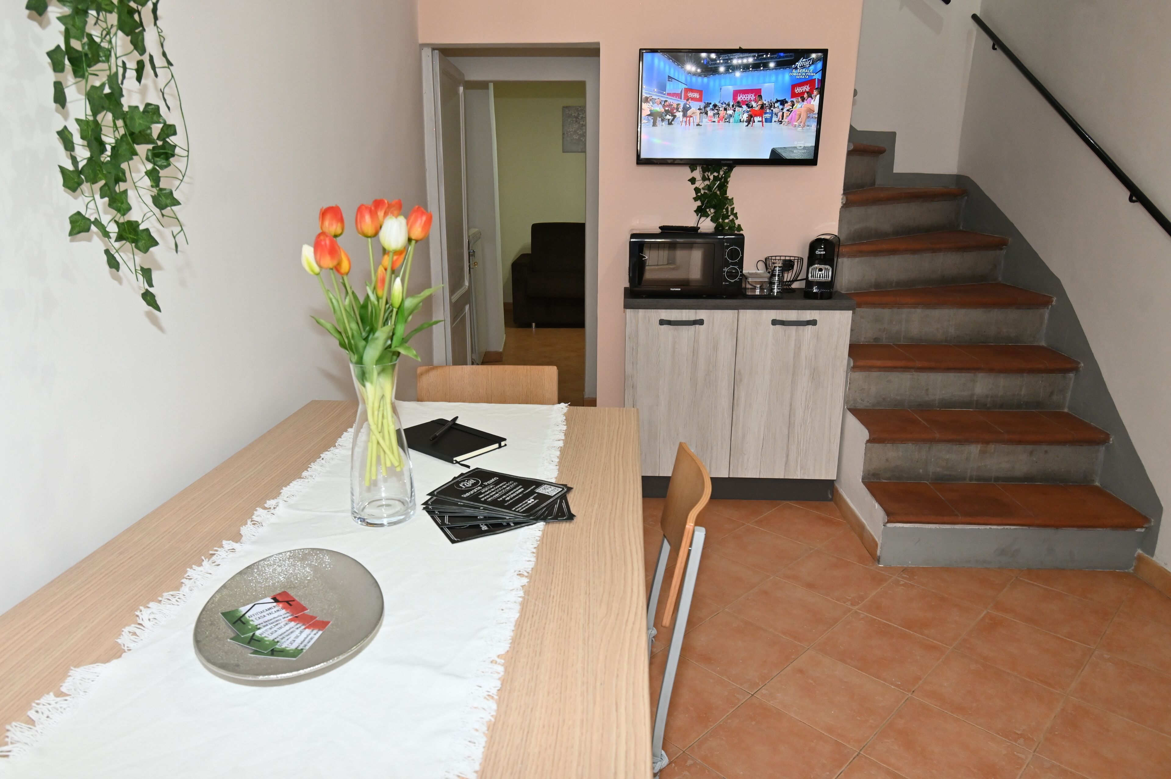 Uma área de refeições com uma mesa posta para dois, um vaso com tulipas, um prato com comida e uma televisão exibindo um evento esportivo.