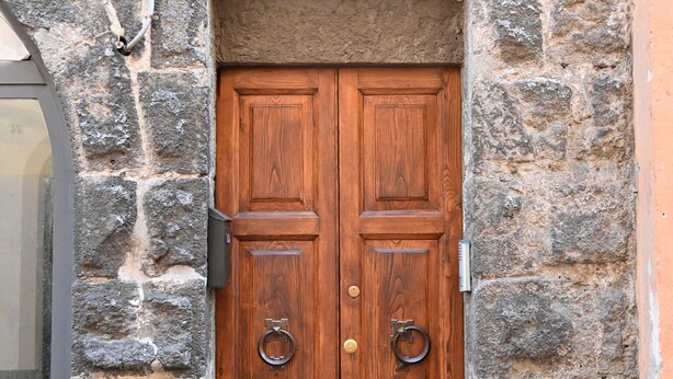 A wooden double door with a metal handle, set in a stone wall with numbered plaques.